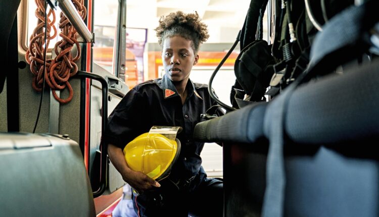 Early 30s woman in station wear with helmet stepping into vehicle and looking away from camera with serious expression.