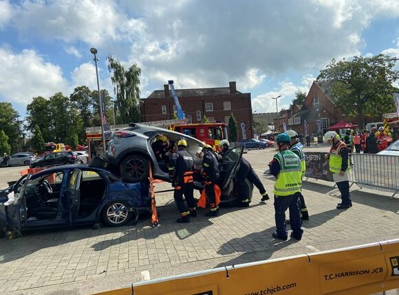 Two cars for demonstration purposes are placed as if they have been in a road traffic accident. Fire Fighters are stood around these cars, some of whom are trying to rescue the 'driver's and passengers. From left to right: Medic Mark Palin, Medic Stephen Fisher, Medic Ashley Capper, Incident Commander Ian Melville, Technical Tom Weate, Technical Simon Moore, Technical David Nelson